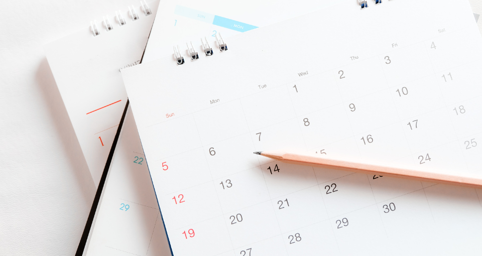 A close-up of a desk with two spiral-bound calendars showing different months and a pencil resting on one of the calendars.