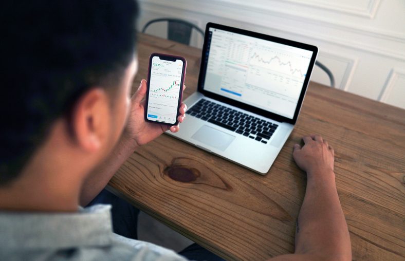 A person is seated at a wooden table, holding a smartphone displaying a data chart, with a laptop in front of them showing similar graphs of data.